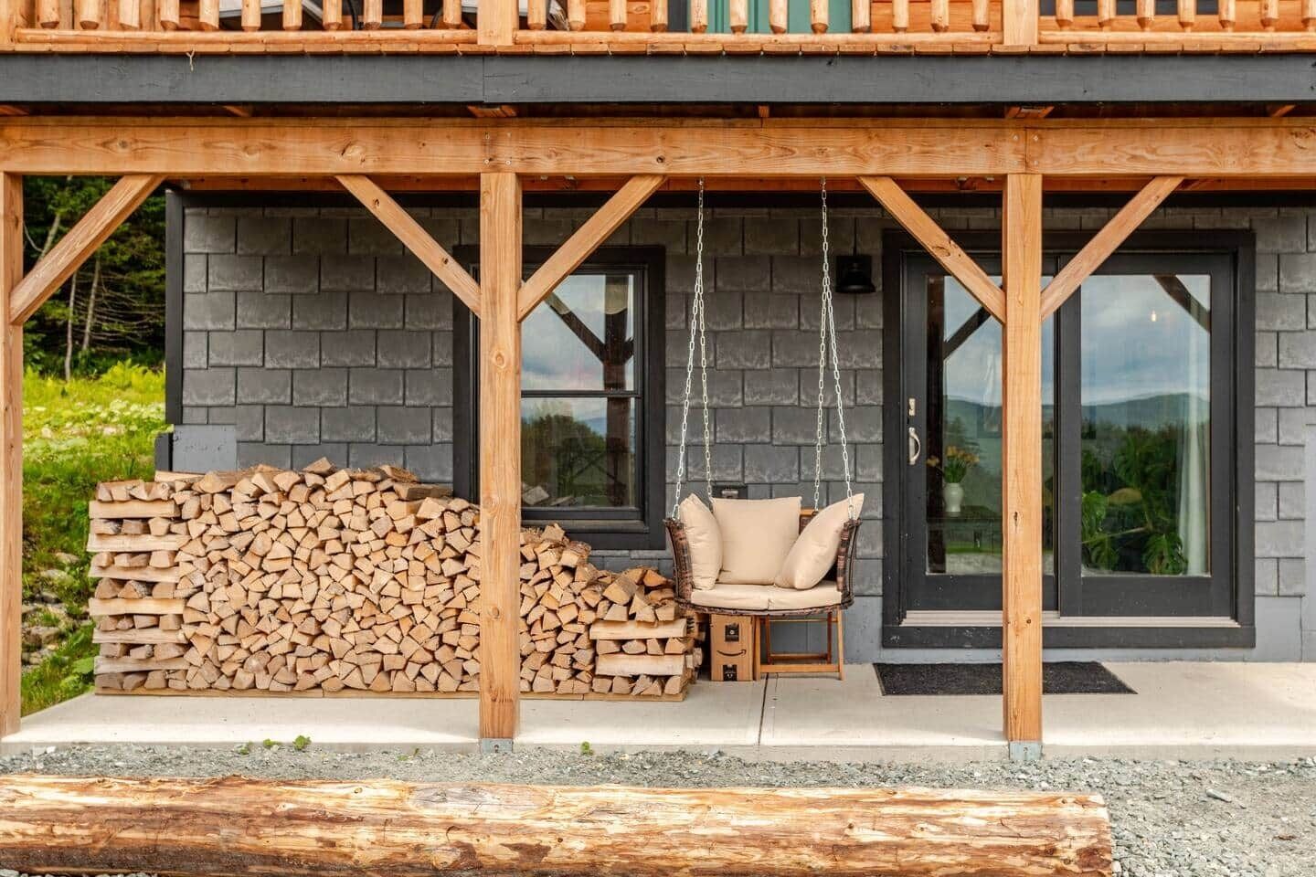 Une terrasse en bois avec un fauteuil suspendu et une pile de bois de chauffage devant un bâtiment doté de grandes fenêtres.
