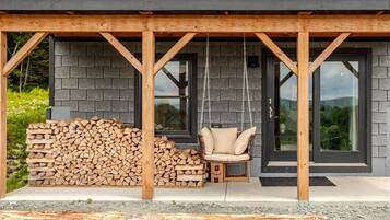 Une terrasse en bois avec un fauteuil suspendu et une pile de bois de chauffage devant un bâtiment doté de grandes fenêtres.