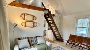 Una habitación estilo loft con techo alto, vigas de madera a la vista, una escalera, un sofá, un banco y un tapete estampado.