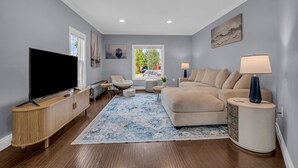A modern living room with a flat-screen TV, a beige sofa, a patterned rug, and a side table with a lamp.