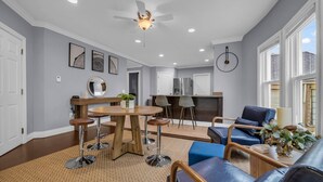A modern living room with a dining area, a kitchen in the background, and large windows.