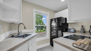 A modern kitchen with white cabinets, stainless steel appliances, a window with a view of greenery, and a built-in oven. - Lighthouse Loft– Chic Getaway in Marine City (Marine City)