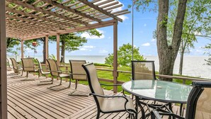 A covered outdoor seating area with wooden deck, chairs, and a table overlooking a body of water.