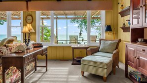 A room with a view of the sea, a desk with books, a wicker chair, and a wooden cabinet.