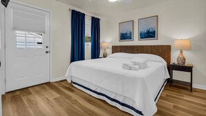 A hotel room with a bed, a ceiling fan, two paintings, a nightstand with a lamp, and a door with a window. - Knot a Care - Lakefront Living with Great Views! (Port Hope)