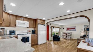 A modern kitchen with wooden cabinets, a white refrigerator, and a stove. There is a dining area with a table and chairs, and a window with a view of the outdoors. - Knot a Care - Lakefront Living with Great Views! (Port Hope)