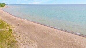 A wide sandy beach with gentle waves, a clear sky, and sparse vegetation. - Steps to the Sand! Ultimate Beach House Get-A-Way! (Port Sanilac)