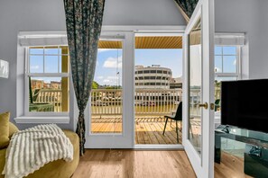 Een kamer met een houten vloer, een gele bank, een witte plaid en een groot raam met uitzicht op een gebouw en een terras.