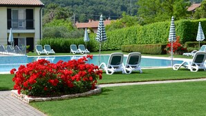 A pool area with lounge chairs and umbrellas, surrounded by greenery and a building in the background.