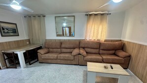 Un salon avec un canapé marron, une table basse en bois, une table à manger avec des chaises, un ventilateur de plafond et un miroir au mur.