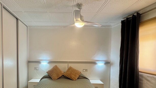Une chambre à coucher comprenant un lit, deux tables de chevet, un ventilateur de plafond et une fenêtre avec des rideaux.