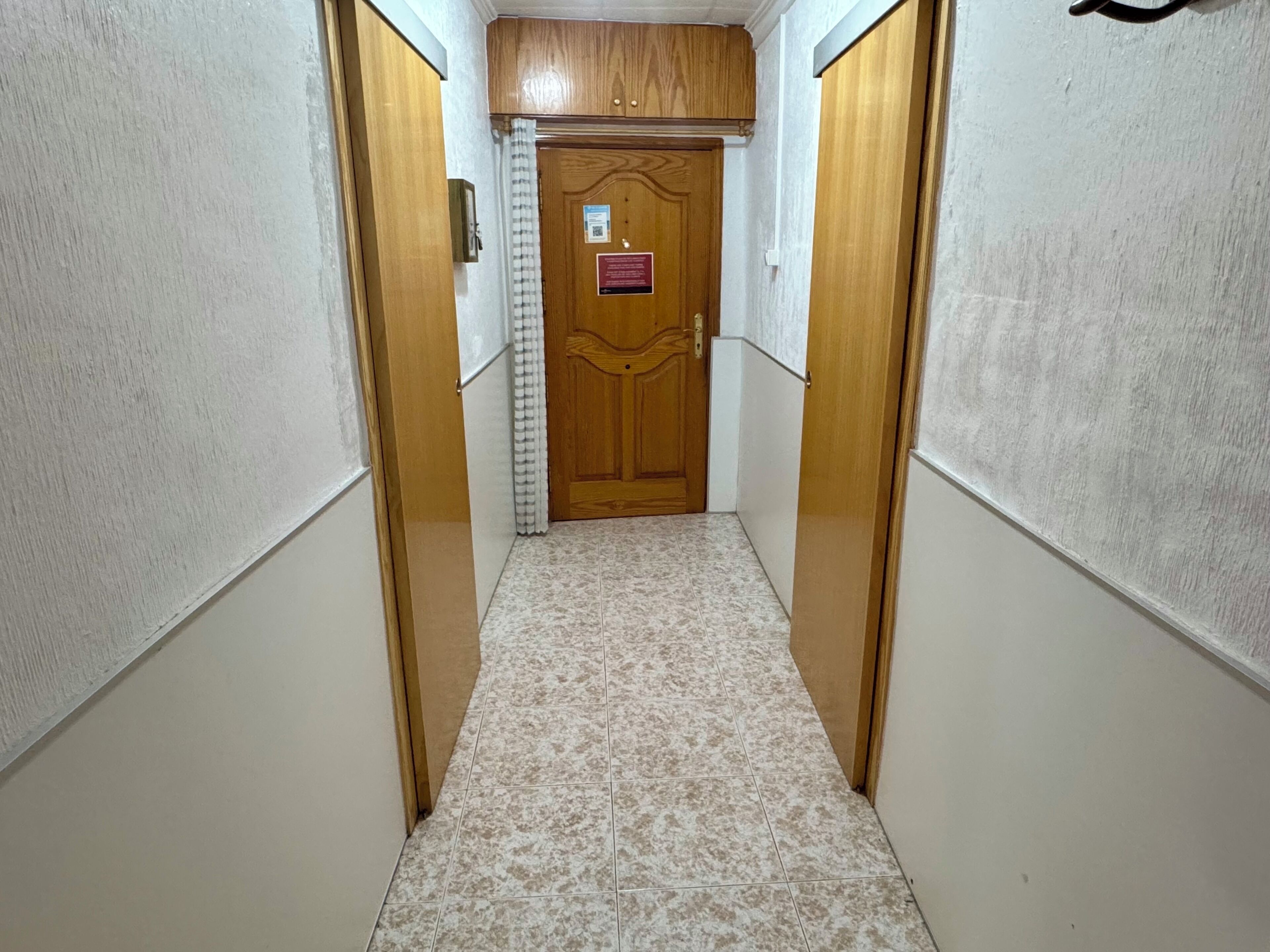 A narrow hallway with a wooden door at the end, white walls, and patterned floor tiles.