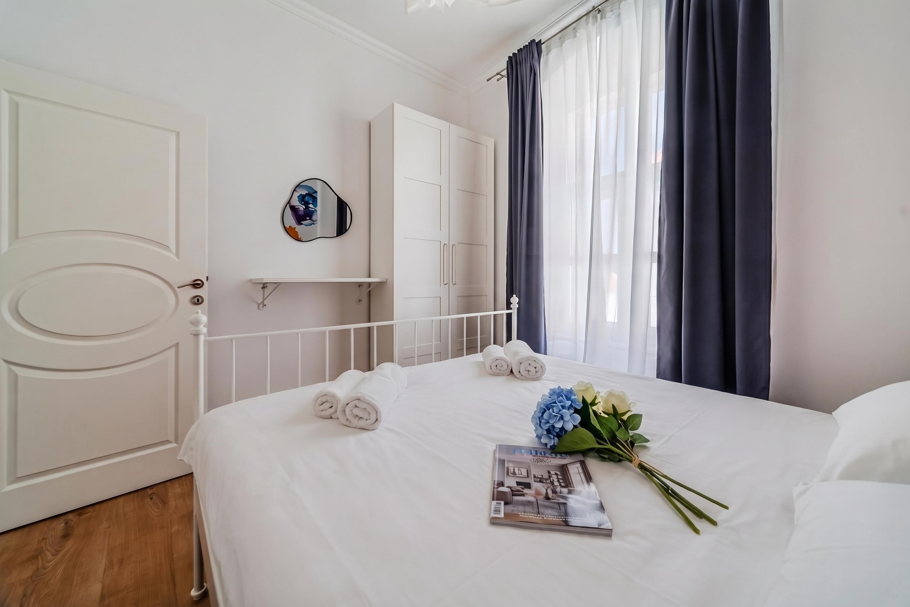A hotel room with a bed, a white door, a white wardrobe, a mirror, and a window with curtains.