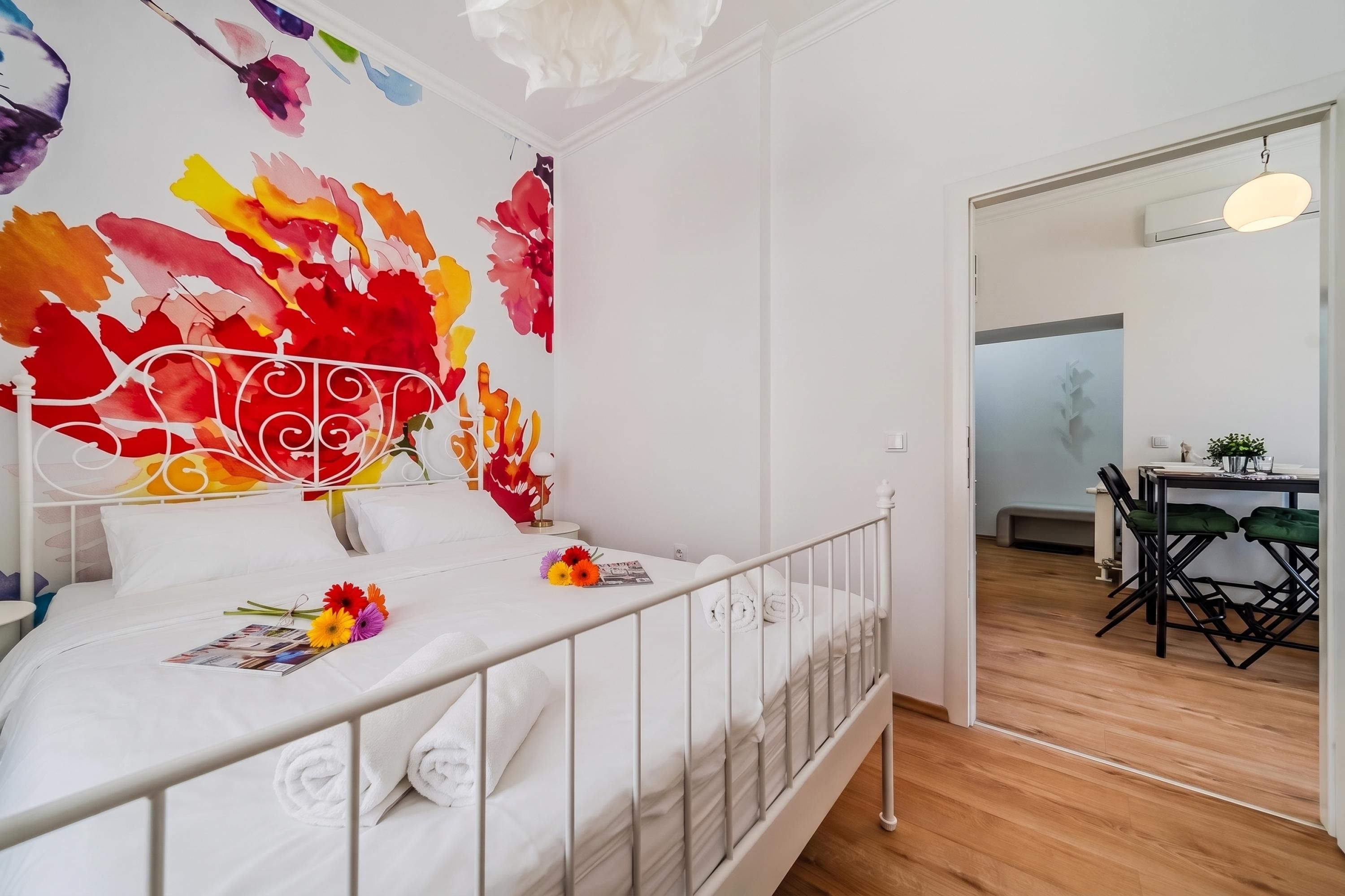 Ein Hotelzimmer mit einem Bett, einem weißen Metall-Kopfteil, einer Wand mit farbenfroher abstrakter Kunst, einem kleinen Tisch mit einer Lampe und einem Essbereich mit Stühlen.
