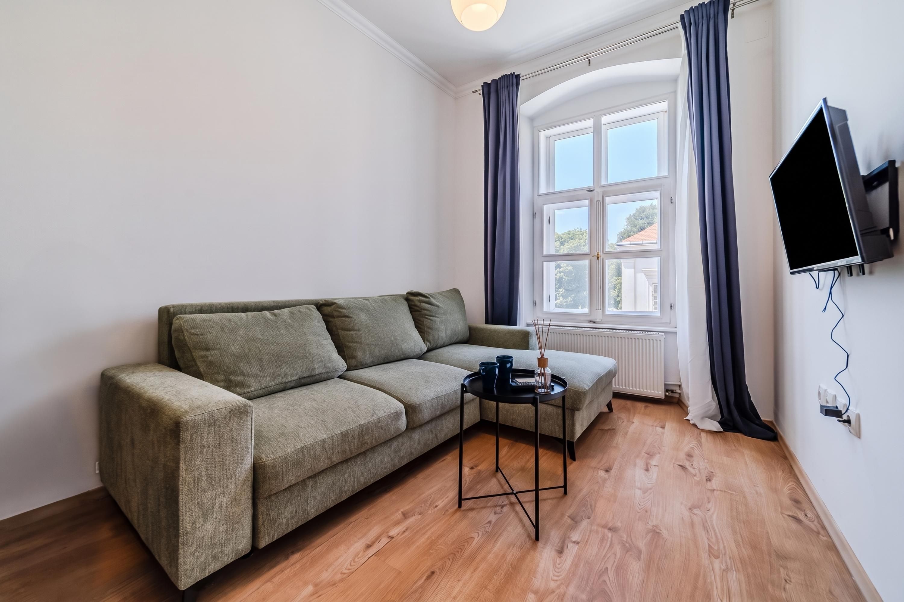 A furnished room with a sofa, a small table, a TV mounted on the wall, and a window with curtains.