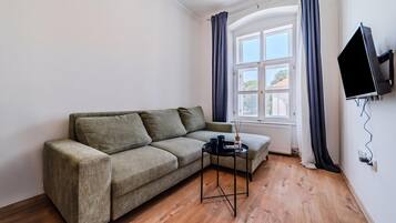 A furnished room with a sofa, a small table, a TV mounted on the wall, and a window with curtains.