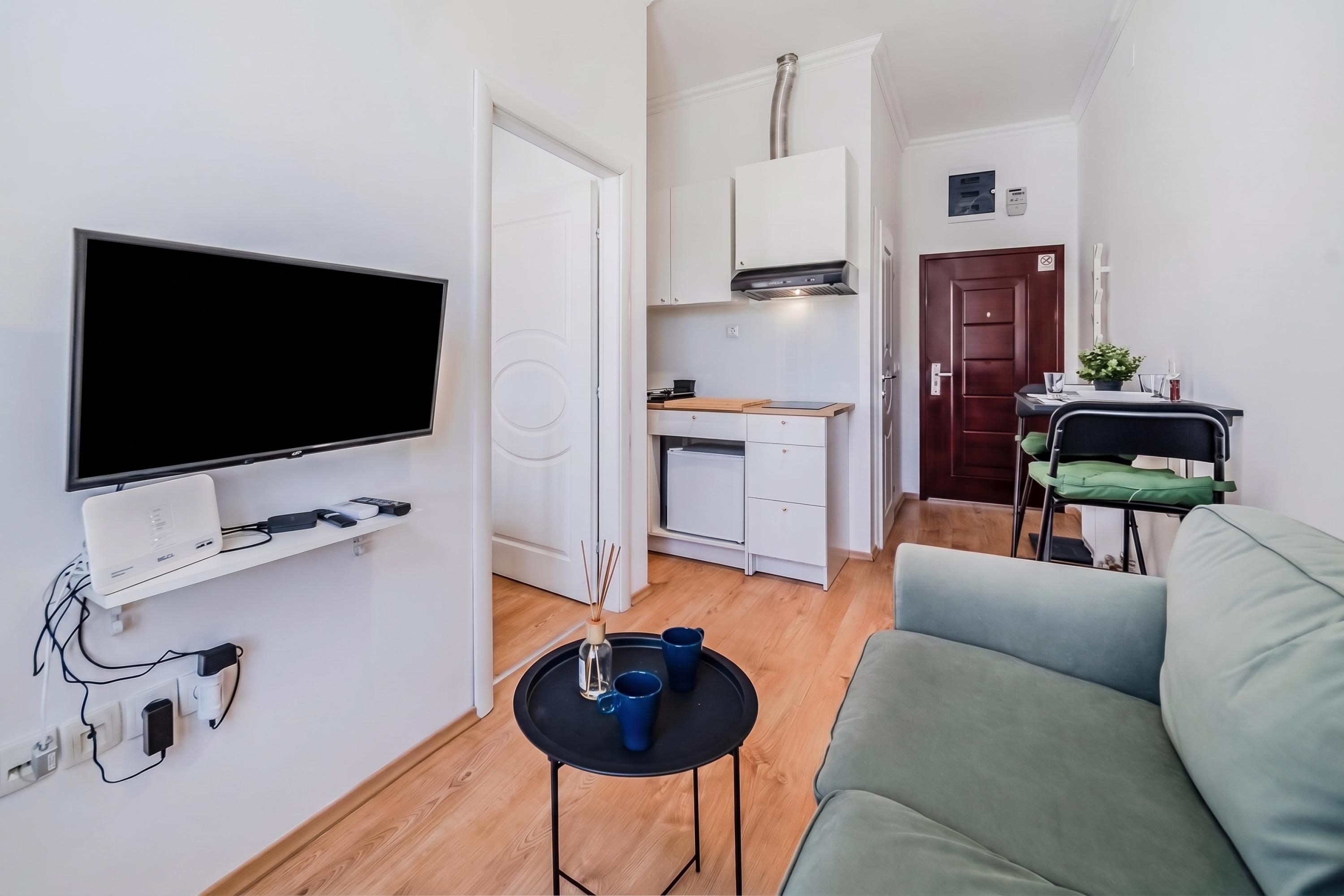 A compact living space with a flat-screen TV, a small table with two cups, a sofa, a kitchenette with a stove, and a door with a sign.