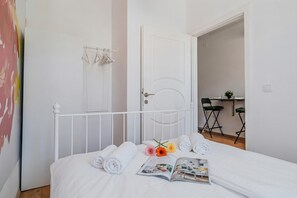 Ein ordentlich bezogenes Bett mit aufgerollten Handtüchern, einer Zeitschrift und Blumen. Eine weiße Tür und ein kleiner Tisch mit einer Pflanze sind ebenfalls zu sehen.