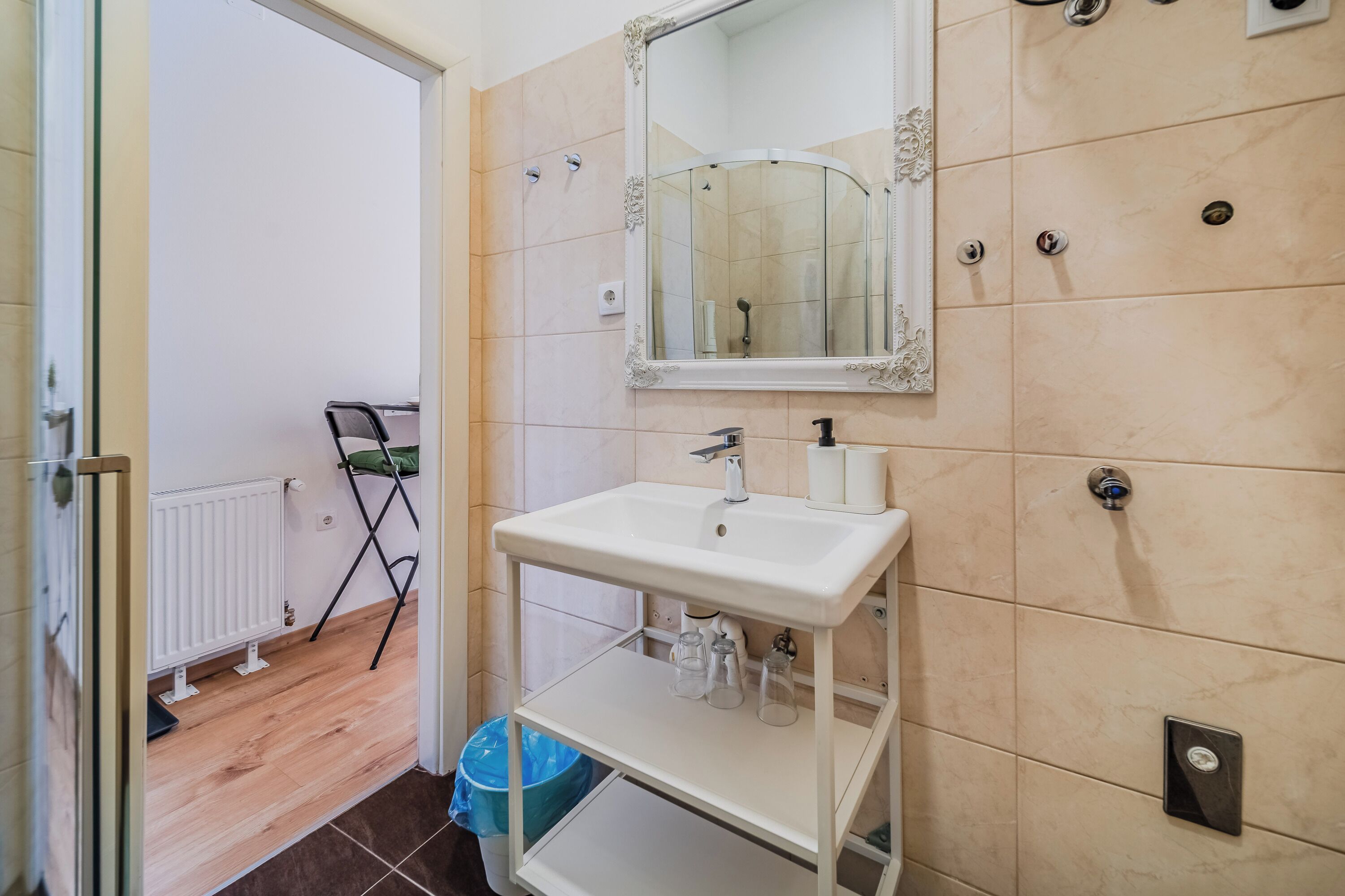 Ein Badezimmer mit einem weißen Waschbecken, Spiegel und gefliesten Wänden. Im Hintergrund ist eine Duschkabine zu sehen.