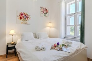 Una cama bien tendida con toallas enrolladas, una revista y flores. Hay dos lámparas de pared y dos cuadros con motivos florales.