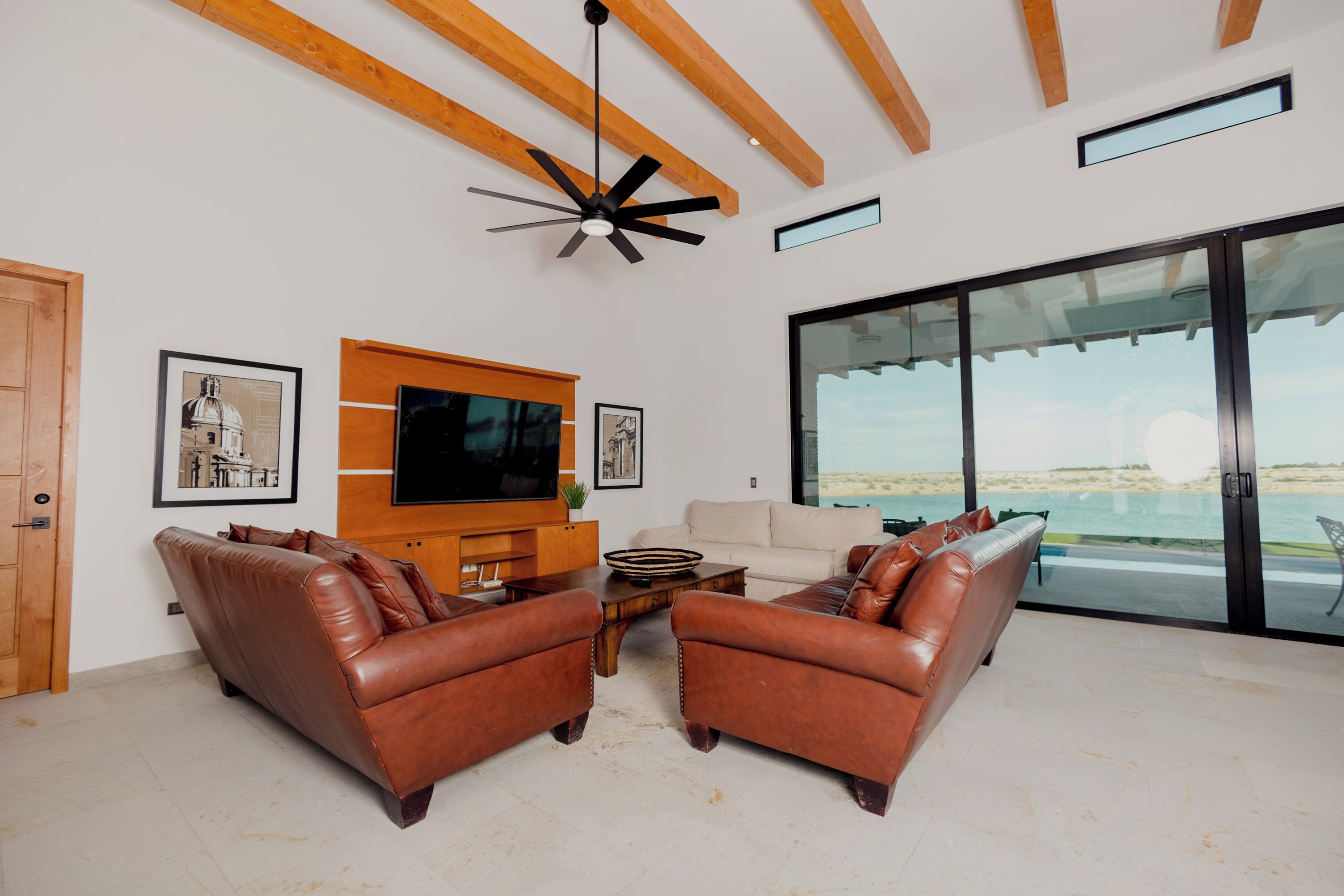 Una sala de estar moderna con sofás de cuero, un televisor de pantalla plana, vigas de madera y un amplio ventanal que da a un balcón con vistas a la playa.