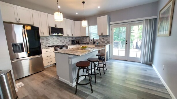 A modern kitchen with white cabinets, a stainless steel refrigerator, a built-in stove, and a large island with bar stools. - Opulent 4 Bdrm, Sleeps 12, Minutes From Downtown! (Cincinnati)