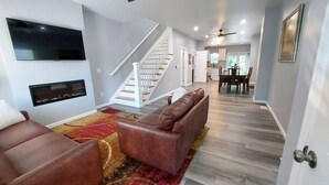 A modern living room with a brown leather sofa, a flat-screen TV mounted on the wall, and a staircase leading to the upper floor. - Opulent 4 Bdrm, Sleeps 12, Minutes From Downtown! (Cincinnati)