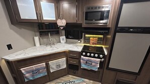 Eine Küche mit Holzausstattung, einer eingebauten Mikrowelle, einem Herd und einem Kühlschrank. Handtücher hängen am Backofen und ein Schild liegt auf der Arbeitsfläche.
