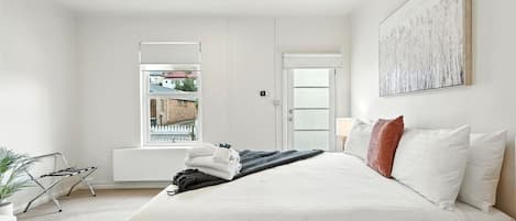 A modern bedroom with a large bed, a view through a window, and a door leading to another room.