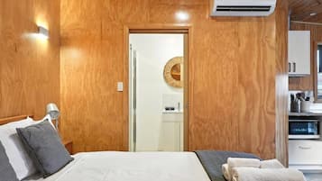 Ein Zimmer mit Holzwandverkleidung, einem Bett, einer Nachttischlampe, einer an der Wand montierten Klimaanlage und einem durchgehenden offenen Durchgang zur Küche.