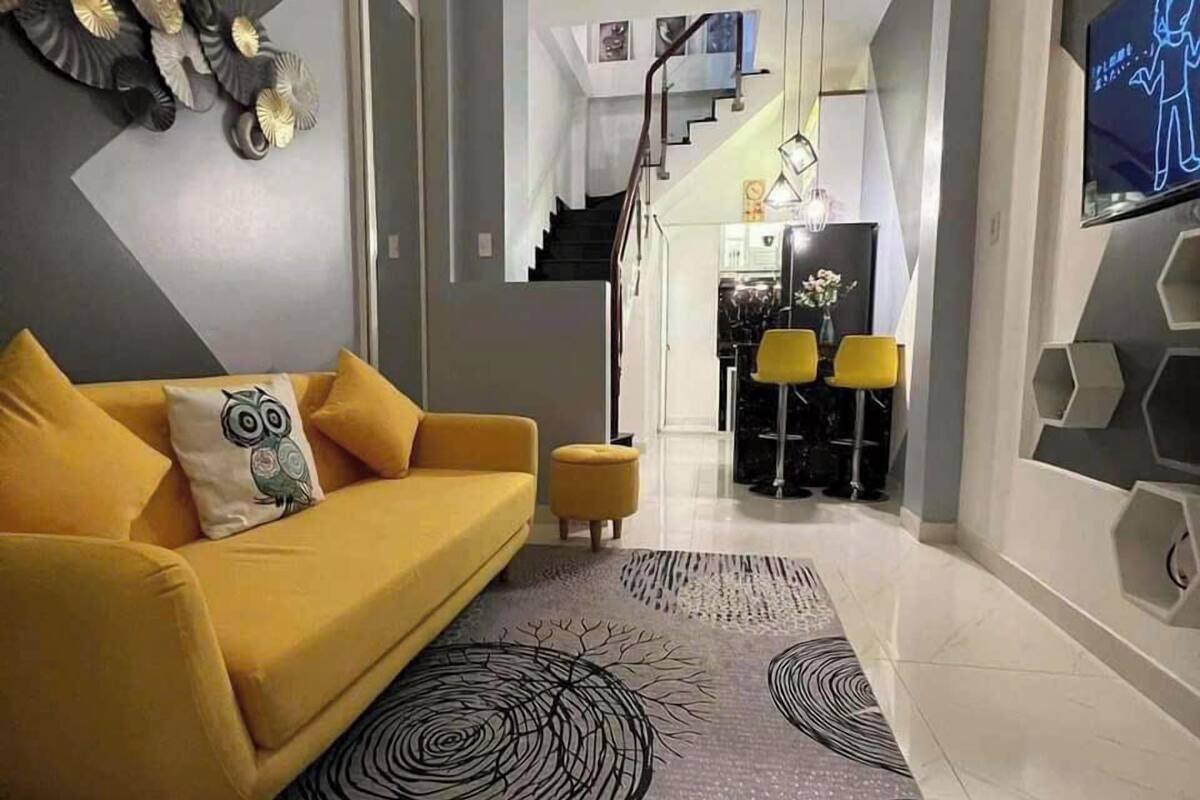 Un salon moderne avec un canapé jaune, un tapis à motifs et un escalier menant à un espace cuisine.
