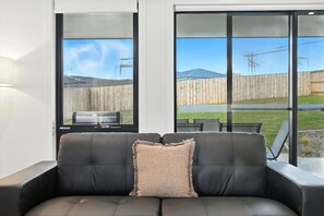 A black leather sofa with a single cushion, positioned in front of a sliding glass door leading to an outdoor patio area with a wooden fence and a grill.