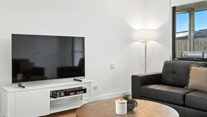 A modern living room with a flat-screen TV, a white entertainment unit, a dark grey sofa, a round wooden coffee table, and a floor lamp. - Modern & Accessible | Old Beach (Old Beach)