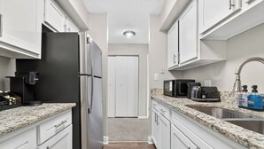 Una cocina estrecha con armarios blancos, un refrigerador de acero inoxidable, una cafetera, un microondas y un tostador.
