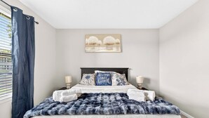 Un dormitorio con una cama, dos lámparas de noche, un edredón blanco y azul mullido, un cojín azul con diseño floral y un cuadro de árboles en la pared.