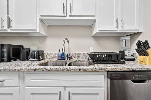 A kitchen with white cabinets, a granite countertop, a stainless steel sink, a microwave, a toaster, a blender, and a knife block.