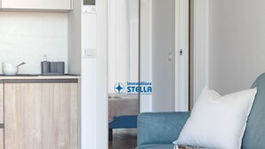 Et moderne køkken med en hvid bordplade, en skål og en elkedel. Der er en blå sofa med en pude og et magasin på. En seng med et hvidt tæppe kan ses i baggrunden.