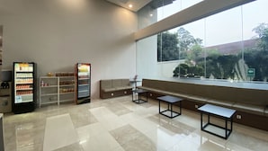 A modern waiting area with a vending machine, seating, and a large window offering a view of greenery and a building. - Padina Soho and Residence (Tangerang)