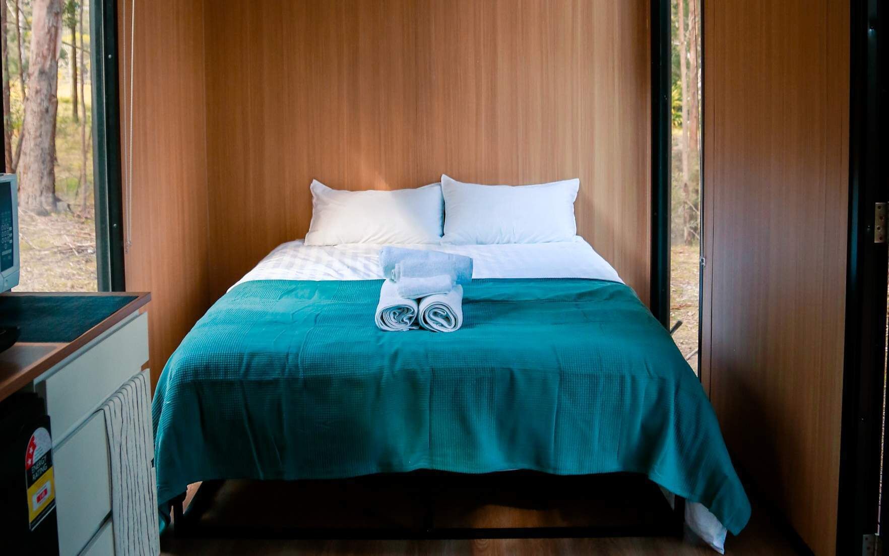 A neatly made bed with white pillows and a teal bedspread, a folded towel, and a small table with a remote control.