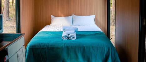 A neatly made bed with white pillows and a teal bedspread, a folded towel, and a small table with a remote control.