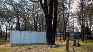 Una estructura grande y rectangular de color azul claro en una zona boscosa, con un sendero de tierra que la lleva hacia ella.
