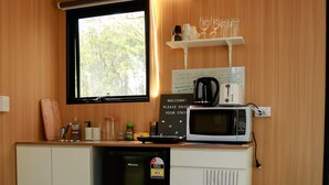 Una kitchenette compacta con microondas, cafetera y un lavamanos pequeño. Hay una ventana con vistas a árboles, y un cartel con instrucciones para usar las instalaciones.