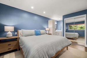 Une chambre à coucher comprenant un lit, des tables de chevet, une commode, un miroir et une fenêtre donnant sur un paysage verdoyant.