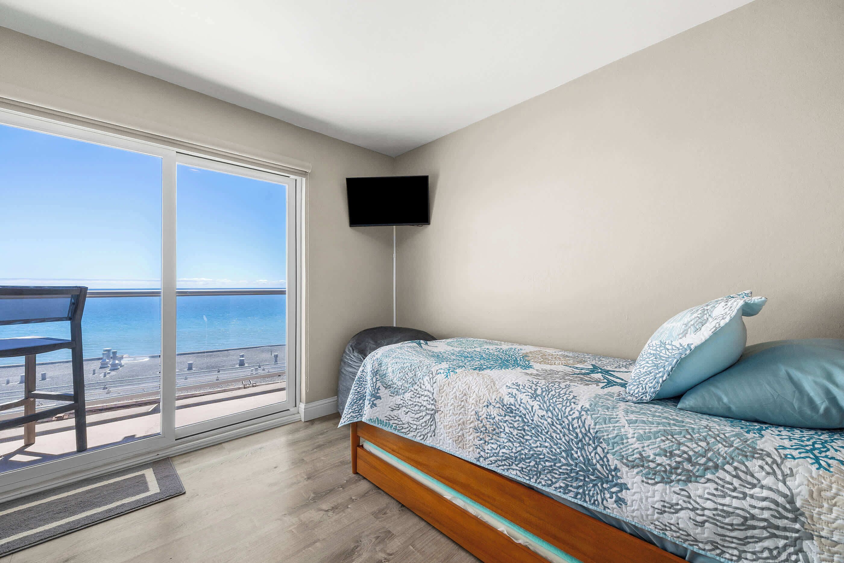 A bedroom with a large window, a bed with patterned bedding, a flat-screen TV mounted on the wall, and a view of the beach.