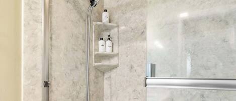 Ducha con cabezal de ducha manual, zona de ducha con cerramiento de vidrio y pared de mármol.