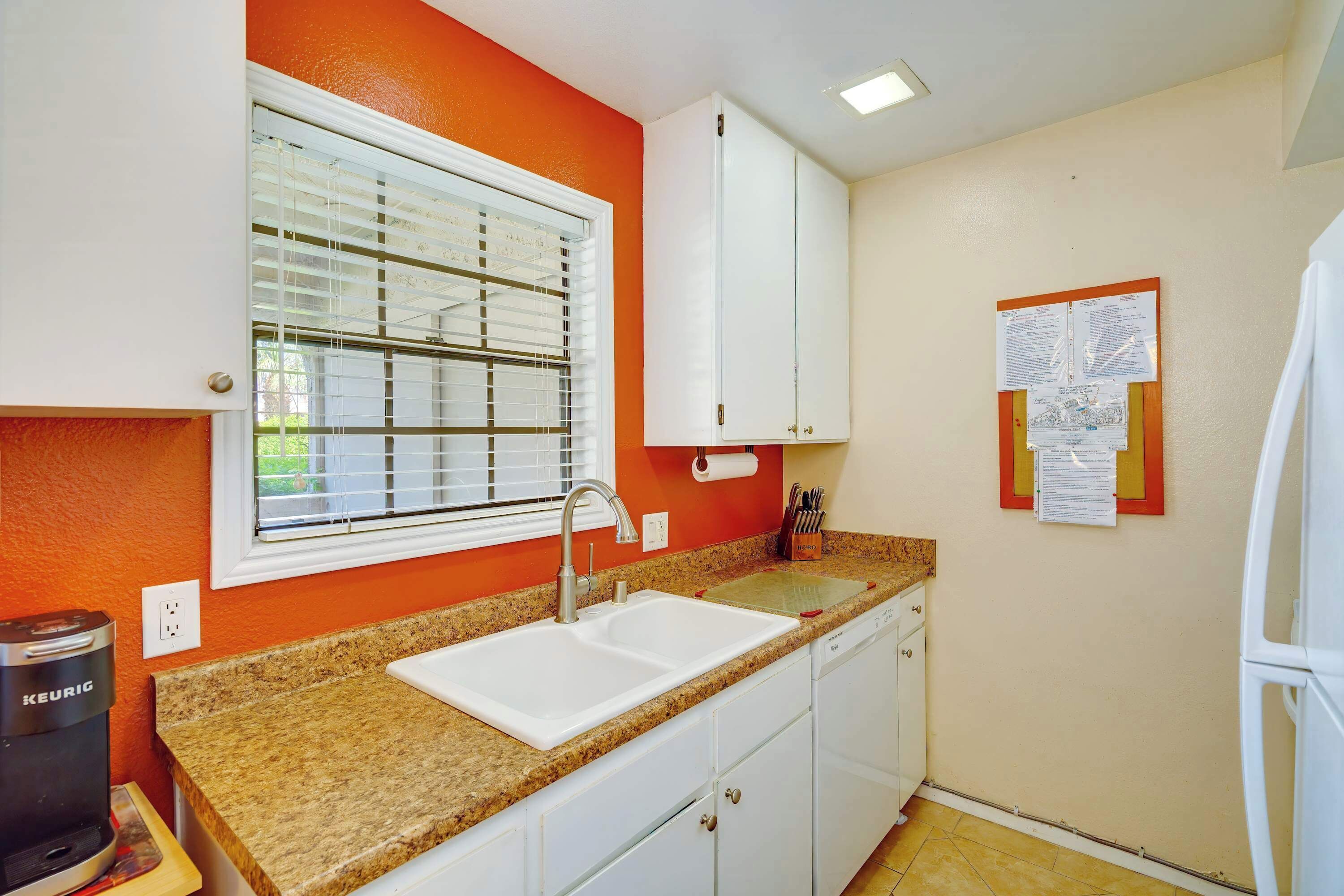 A kitchen with white cabinets, a granite countertop, a built-in sink, a window with blinds, and a coffee maker.