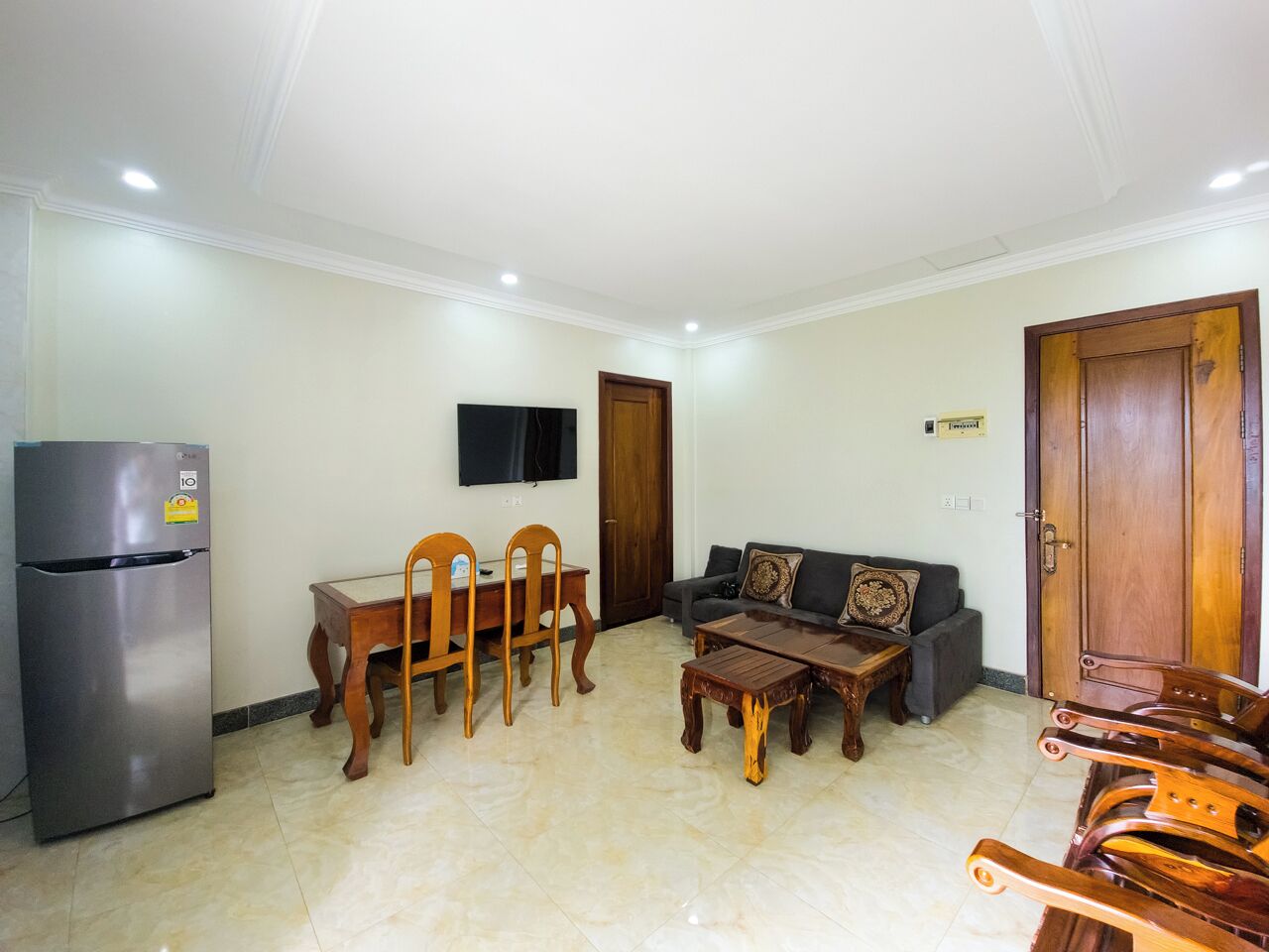 A living room with a dining table, chairs, a sofa, a coffee table, a TV mounted on the wall, and a wooden door.