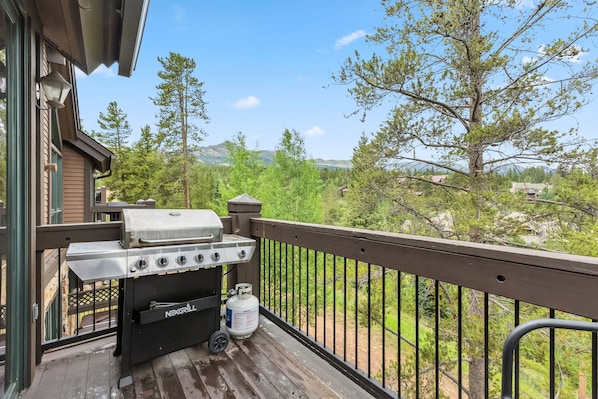 A balcony with a grill, a propane tank, and a view of a forest and mountains.