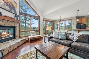 A spacious living room with a stone fireplace, large windows, a leather sofa, and a wooden coffee table. - Snowflake Lift Access | Hot Tub + Views (Breckenridge)