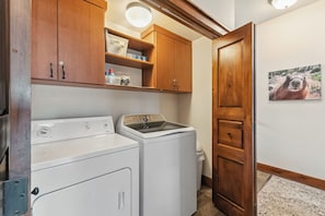 A laundry room with two washers and dryers, wooden cabinets, a shelf with toiletries, and a door with a framed picture.